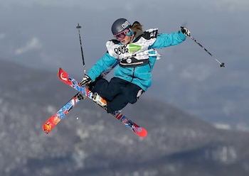 Esquiador en el aire con chaqueta azul, casco, gafas y bastones de esquí. Lleva esquís coloridos y un dorsal con el texto 'Dew Tour'