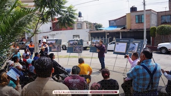 Grupo de personas sentadas y de pie en la calle, escuchando a un hombre que señala paneles informativos del Trolebús Ixtapaluca. Hay árboles y casas