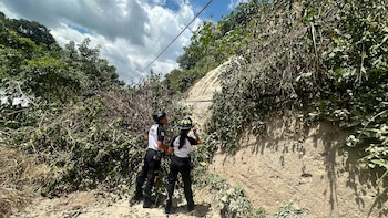 Dos personas con uniformes de bomberos y cascos inspeccionan una gran pila de ramas y hojas caídas junto a una ladera de tierra, bajo cables eléctricos y un cielo nublado