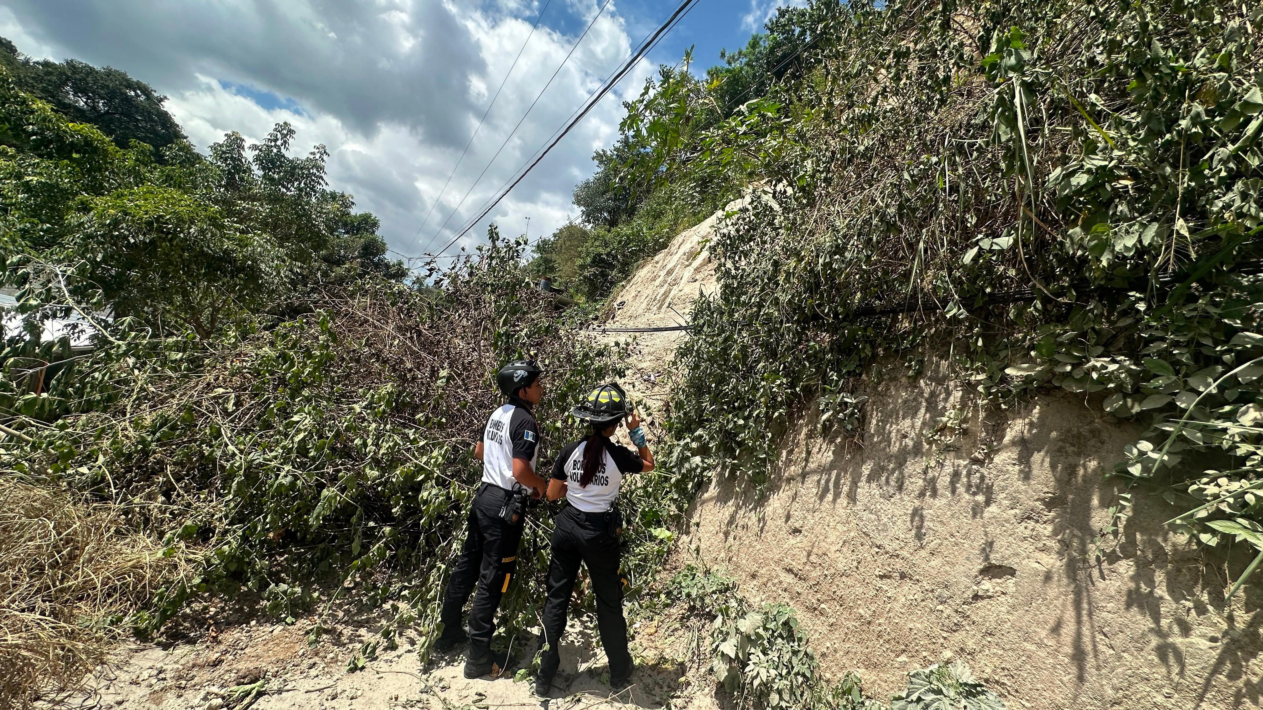 Dos bomberos voluntarios de Guatemala revisan la acumulación de tierra y vegetación caída en un camino rural, con postes y cableado eléctrico visibles al fondo. (Bomberos Voluntarios de Guatemala)