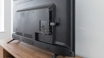 Vista trasera de un televisor negro sin cables visibles, con un pequeño adaptador HDMI inalámbrico conectado a un puerto lateral sobre un mueble de madera.