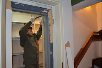 Un hombre con guantes y gorra utiliza una herramienta en el marco de una puerta de madera junto a una escalera