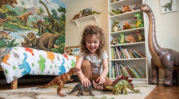 Una niña sobre una alfombra manipula tres figuras de dinosaurios en una habitación con peluches, libros y un mural de dinosaurios.