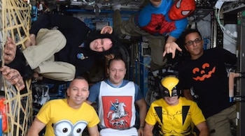 Seis personas en el interior de una estación espacial visten camisetas estampadas, disfraces y máscaras mientras flotan en el habitáculo