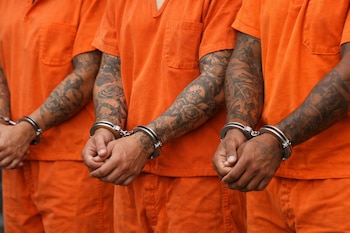 Tres personas con uniformes de prisión naranjas. Sus brazos tienen tatuajes y sus manos están aseguradas con esposas de metal.