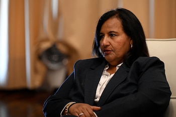 Mujer con cabello oscuro, traje oscuro, camisa blanca, sentada en silla clara, manos entrelazadas. Fondo de cortinas beige