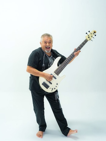 Hombre canoso con barba y bigote, vestido con camiseta negra y pantalón negro, sostiene un bajo eléctrico blanco sobre un fondo blanco. Está descalzo