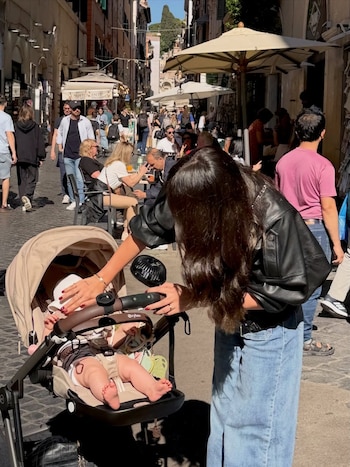 Una mujer de cabello oscuro atiende a un bebé en un cochecito beige con sombrero blanco, en una calle concurrida con restaurantes y gente