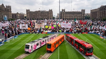 Vista aérea de una multitud manifestándose en una plaza histórica con edificios antiguos, mientras dos trenes y un autobús rojo atraviesan el primer plano.