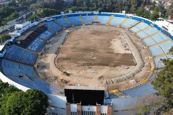 Vista aérea de un estadio vacío con el campo de tierra, gradas azules y amarillas, excavadoras, una pantalla gigante y la palabra GUATEMALA en los asientos