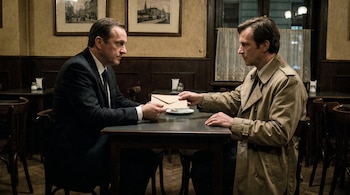 Dos hombres sentados frente a frente en una mesa de un café, uno con traje oscuro y el otro con gabardina, están intercambiando un sobre beige.
