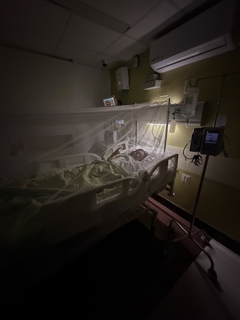 Una mujer recostada en una cama de hospital cubierta por un mosquitero blanco, rodeada de equipos de monitoreo médico y bolsas de suero