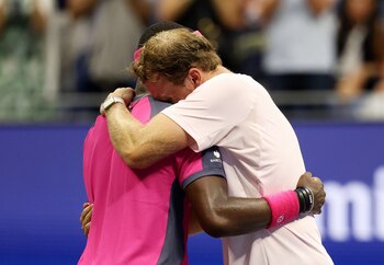 Dos jugadores de tenis se abrazan en la pista; uno viste de color rosa intenso y el otro lleva una camiseta rosa claro