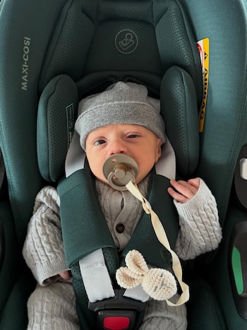 Un bebé con gorro gris y suéter tejido está sentado en una silla de auto verde oscura, con chupete y un sujetador de chupete con forma de conejito