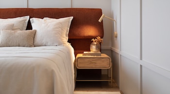 Cama con cabecero tapizado, almohadas, sábanas blancas, mesita de noche de madera, lámpara de pared dorada, jarrón con flores secas y libros.