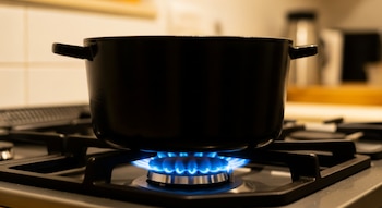 Una olla negra descansa sobre una hornalla encendida de una cocina a gas, con una brillante llama azul visible debajo. El fondo está cálido y desenfocado.