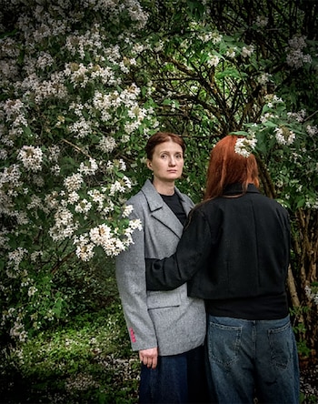 Una mujer con chaqueta gris mira al frente mientras otra de espaldas la abraza junto a plantas florecidas