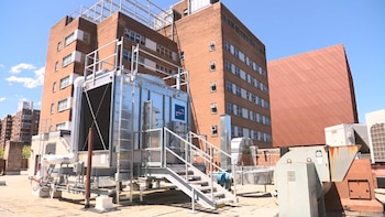 Equipo de climatización industrial en una azotea con escaleras y tuberías; al fondo, varios edificios de ladrillo con ventanas y un cielo azul con nubes