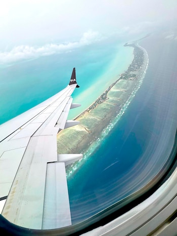 Vista desde la ventanilla de un avión que muestra parte del ala con el logo "FIJI", una isla alargada con edificaciones, agua turquesa y azul, y nubes