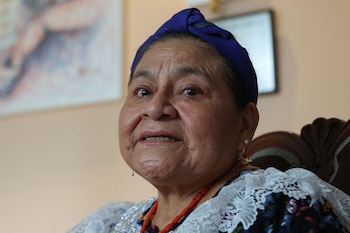 Primer plano de Rigoberta Menchú con turbante azul, blusa blanca con encaje, collar rojo de cuentas y aretes de perla. Fondo beige con cuadro borroso