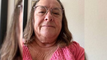 Primer plano de una persona con arrugas, cabello largo castaño, gafas de montura y camiseta rosa sobre un fondo liso