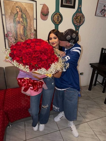 Uma jovem com um buquê de flores vermelhas é beijada na bochecha por um homem. Ao fundo, imagem religiosa e dois cintos de boxe