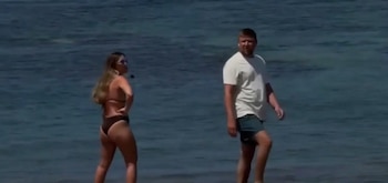 Dos personas, un hombre y una mujer, de pie en la orilla de una playa con agua de mar de color azul oscuro al fondo