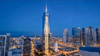 Un rascacielos alto y moderno con la palabra 'TRUMP' y un diseño de bandera estadounidense, iluminado en una ciudad al anochecer bajo un cielo azul oscuro