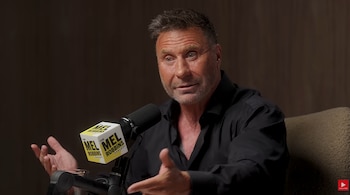 Un hombre con camisa negra gesticula frente a un micrófono con el logotipo de The Mel Robbins Podcast en una sala con fondo oscuro