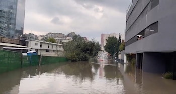 Una calle inundada con un coche, un rascacielos de cristal, un edificio gris, un baño portátil azul, edificios blancos y un cielo cubierto de nubes