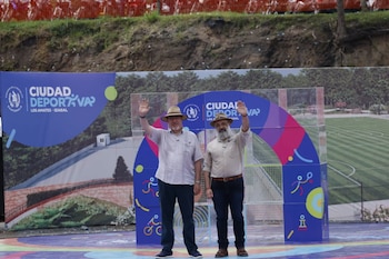 Dos hombres con sombrero levantan la mano frente a un panel publicitario con la inscripción Ciudad Deportiva Los Amates