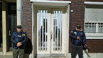 Dos agentes uniformados de la PFA, uno con escopeta, custodian un edificio de ladrillo marrón con puerta doble blanca. Varias personas se ven detrás del cristal