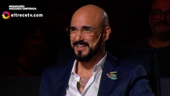 Hombre calvo con anteojos, barba, saco azul y camisa blanca con un micrófono de diadema sobre un fondo oscuro