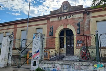 Fachada de una escuela antigua de ladrillo en tonos beige y rojizo con la inscripción 'E.N.E.T. N°1' sobre una entrada arqueada, rodeada por una reja