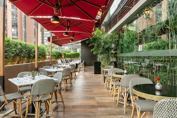 Patio exterior de restaurante con mesas y sillas de mimbre bajo grandes sombrillas rojas. Hay plantas verdes, un suelo de madera y ventanas de un edificio al fondo