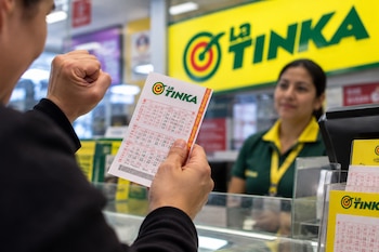 Unas manos sostienen un boleto de lotería de La Tinka frente a un mostrador con el logotipo del juego y una vendedora al fondo