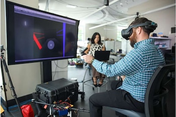 Un hombre con barba usa una computadora portátil frente a una pantalla grande con gráficos digitales tridimensionales