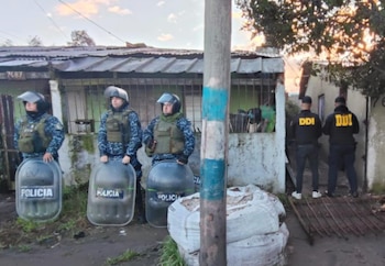 Tres policías antidisturbios con escudos y dos agentes de DDI con chalecos negros frente a una vivienda modesta. Cielo claro al atardecer
