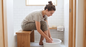 Una mujer sentada en un banco de madera lava sus pies en un recipiente blanco sobre el suelo azulejado de un baño.