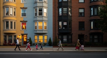 Dos edificios de departamentos enfrentados en Boston al atardecer; uno con ventanas iluminadas y otro con carteles de 'For Rent'. Personas caminan por la acera.