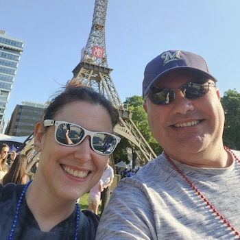 Un hombre con gorra y gafas de sol sonríe junto a una mujer con gafas de sol frente a una torre de estructura metálica