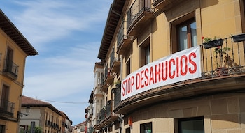 Un edificio de piedra con múltiples balcones y una pancarta blanca con el texto rojo 'STOP DESAHUCIOS' colgada de un balcón superior, bajo un cielo azul claro.