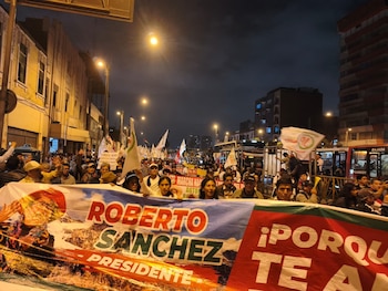 Multitud de personas en una calle urbana por la noche. Llevan pancartas con el nombre Roberto Sanchez y banderas. Se observan edificios y autobuses