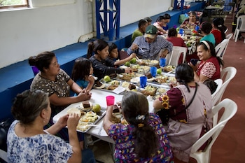 Varias personas, incluyendo hombres, mujeres y niños, sentadas en mesas largas comiendo de platos con comida variada y bebidas en vasos de plástico