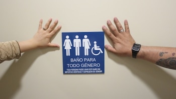 Dos manos, una con chaqueta beige y anillo, otra con reloj y tatuajes, flanquean un letrero azul de "Baño para Todo Género" en una pared clara