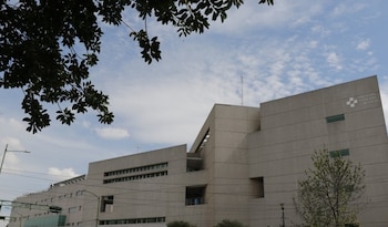 Edificio de color claro con ventanas y el logo del Hospital General de México en la fachada, ramas de árbol con hojas verdes en la parte superior, cielo con nubes