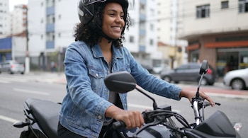 Mujer joven de piel morena con cabello rizado, casco negro, chaqueta de mezclilla azul, sentada en una motocicleta negra en una calle urbana.