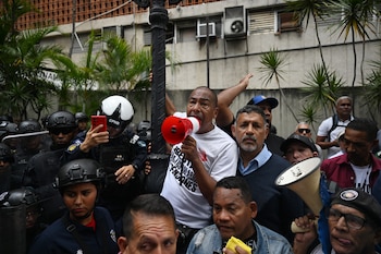Multitud de personas con varios hombres sosteniendo megáfonos, rodeados por agentes de policía con cascos negros y escudos. Palmeras y edificios visibles