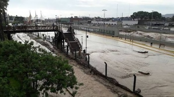 Agua turbia y lodo cubren una vía vehicular rápida junto a un puente peatonal de madera, barandas y árboles
