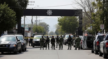 Soldados y policías con vehículos oficiales en una calle con cinta de seguridad, frente a una estructura negra con un emblema, árboles a los lados.